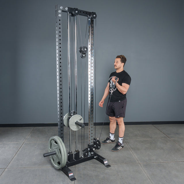 A man stands by a tall, black cable machine in a gym with gray walls and a concrete floor, using Bells of Steel Fat Bar - Single D Handle attachments to enhance grip strength.