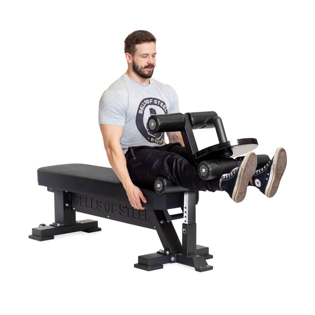A man uses the Bells of Steel Hero Heavy-Duty Weight Bench with a leg extension machine attachment, lifting padded rollers with his legs.