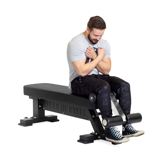 A man in a gray t-shirt and black pants sits on the Bells of Steel Hero Heavy-Duty Weight Bench, arms crossed as he performs a sit-up with his feet tucked under the padded rollers.