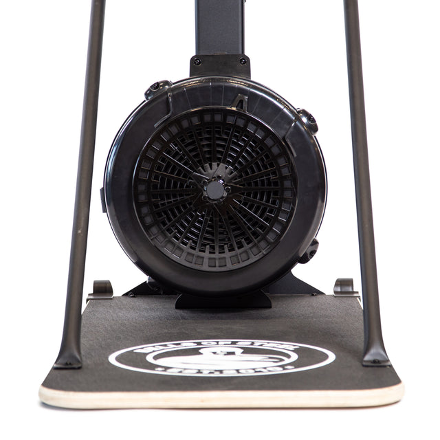 Close-up of the Bells of Steel Blitz Ski Trainer, showing the fan and base with an air-powered flywheel, circular vent, and logo on the platform. Two support legs frame this black cross-country ski workout device.