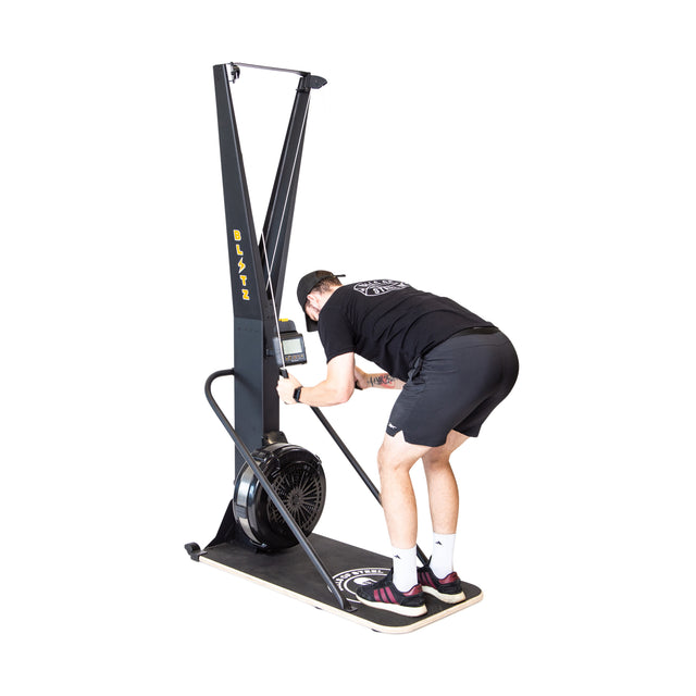 Wearing athletic gear, a person uses the Bells of Steel Blitz Ski Trainer with its air-powered flywheel, pulling handles downward while leaning forward on a black platform against a white background, simulating cross-country skiing.