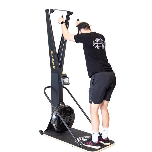 Wearing athletic clothes, a person uses the Bells of Steel Blitz Ski Trainer, gripping the handles and leaning forward to mimic cross-country skiing indoors for a full-body workout.