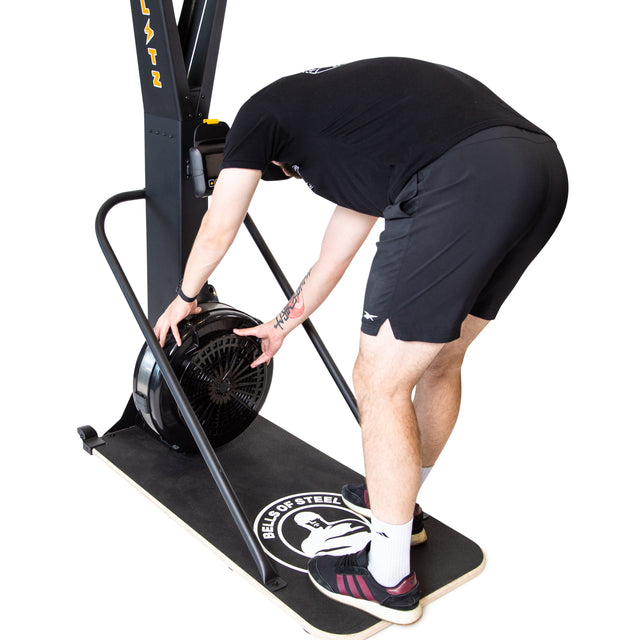 Wearing athletic wear, a person bends forward to adjust the air-powered flywheel of the Bells of Steel Blitz Ski Trainer, which sits on a black platform against an isolated white background.