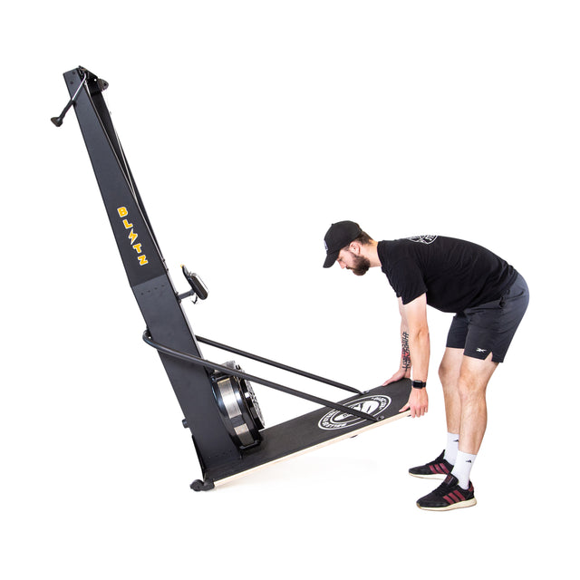 A man in athletic wear tilts the Bells of Steel Blitz Ski Trainer, which features an air-powered flywheel base, preparing to move or store it on a white background.