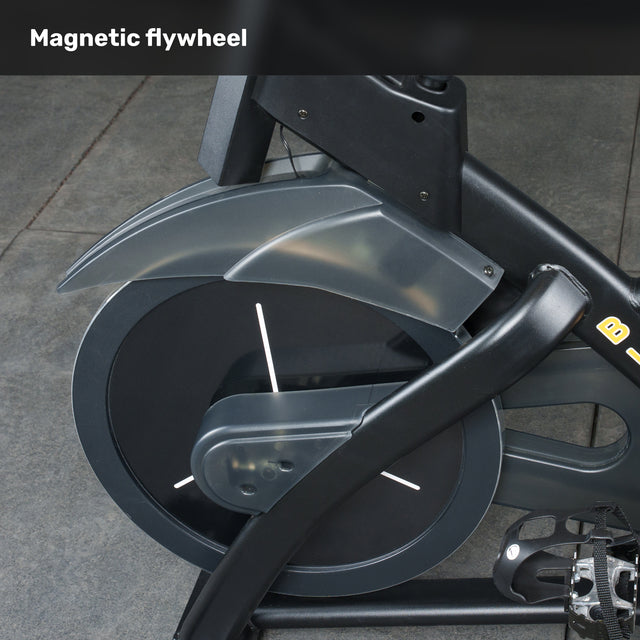 Close-up of the Bells of Steel Blitz Indoor Cycling Bike’s magnetic flywheel, showcasing black and gray parts with metal pedals on a stone-tiled floor. "Magnetic resistance" appears at the top.