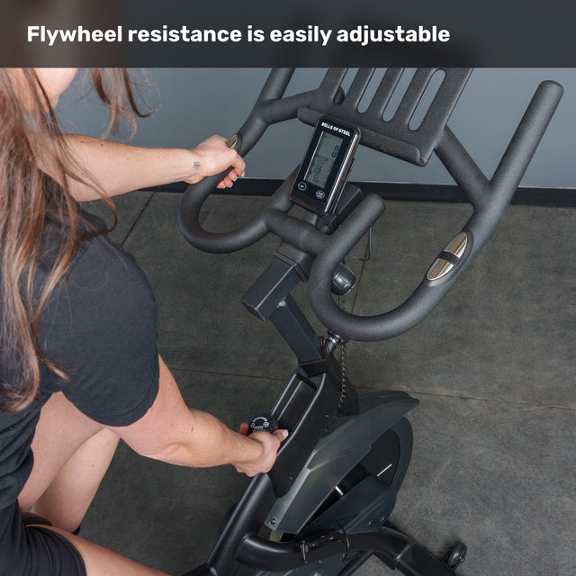 A person adjusts the magnetic resistance knob on the Bells of Steel Blitz Indoor Cycling Bike. The display screen and adjustable handlebars are visible. Text at the top reads, "Flywheel resistance is easily adjustable.