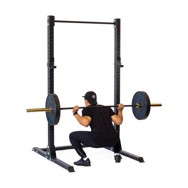 A person in black athletic wear and a cap is performing a barbell squat using the Bells of Steel Hydra Squat Stand - Prebuilt (3" x 3", ⅝" Holes), with the barbell resting across their shoulders.
