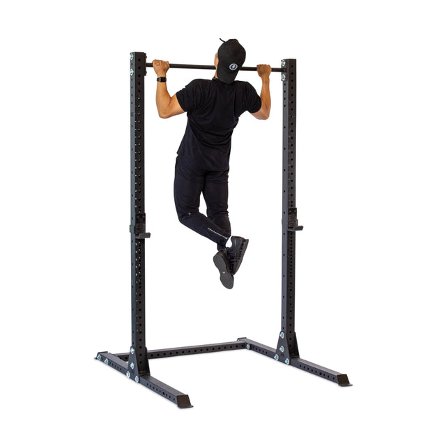 A man in black athletic wear and a cap performs a pull-up on the Bells of Steel Hydra Squat Stand - Prebuilt (3" x 3", ⅝" Holes), set against a white background—an essential addition to any home gym.
