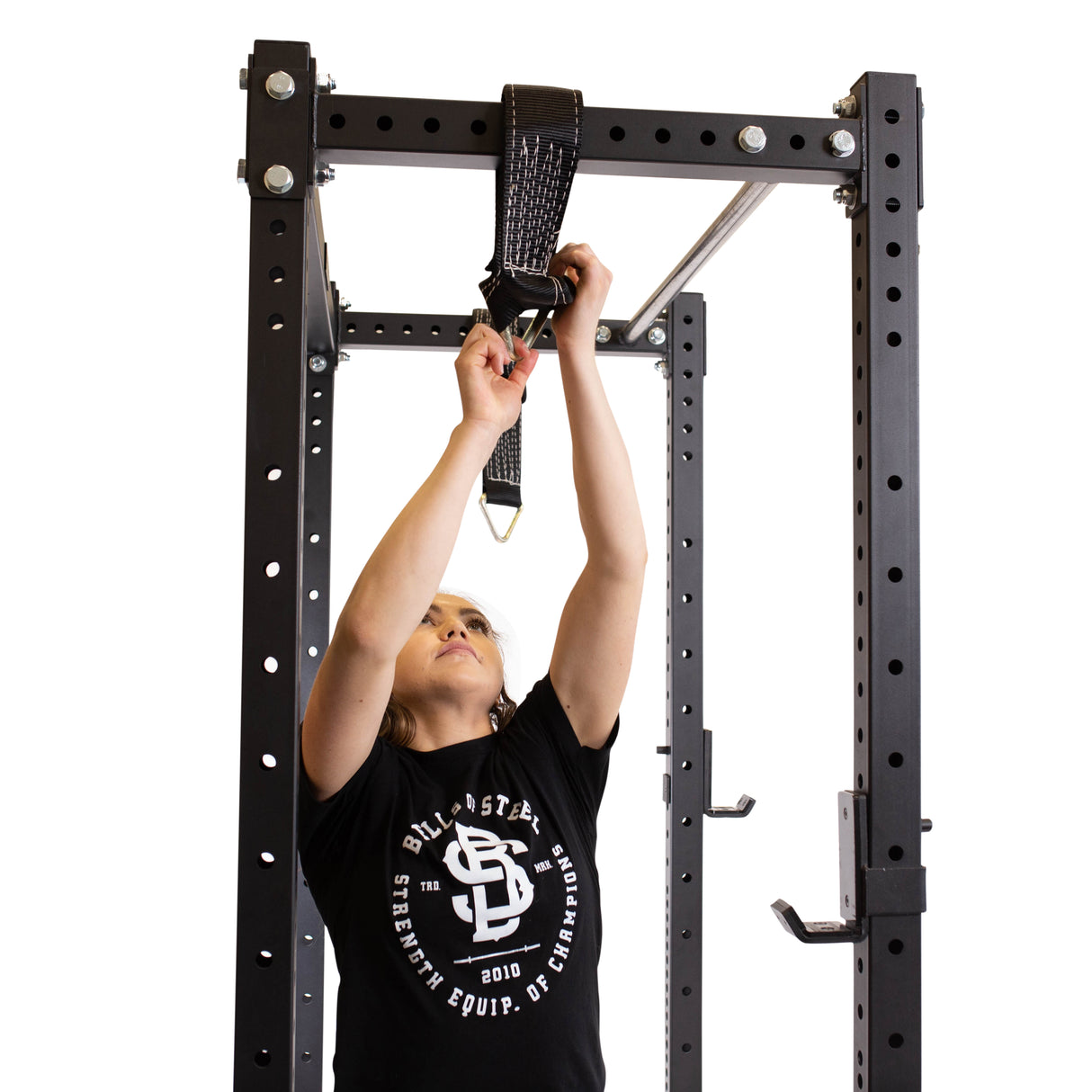 A person in a black shirt adjusts the strap of a Bells of Steel Suspension Spotter Straps And Bodyweight System on a squat rack with pull-up bar attached, skillfully configuring safety spotter straps. The metal frame, featuring evenly spaced holes for adjustments, sits against a white background.