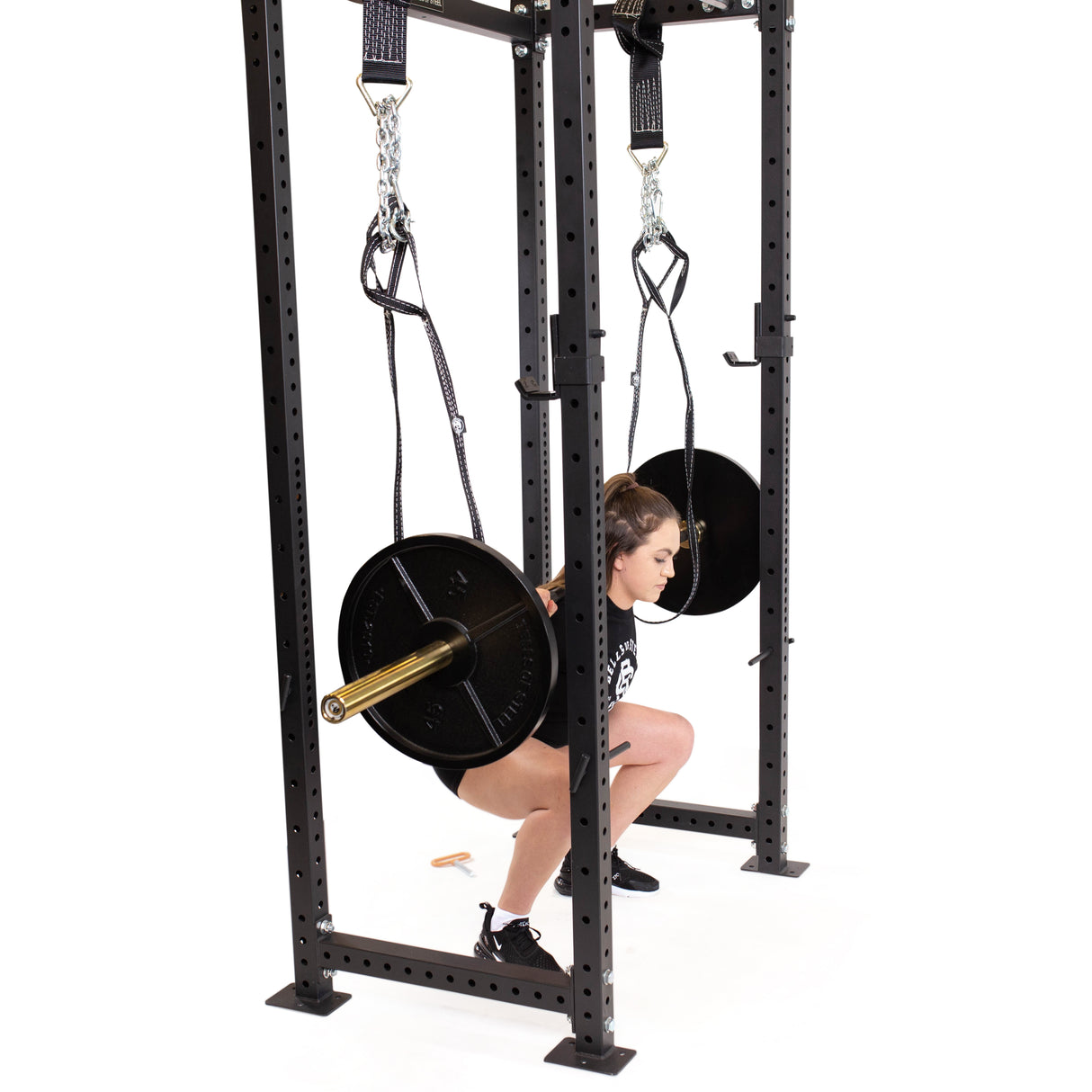 A person uses the Bells of Steel Suspension Spotter Straps and Bodyweight System to squat under a weighted barbell. This system, ideal for home gyms, offers stability and assistance during squats with its metal frame support.