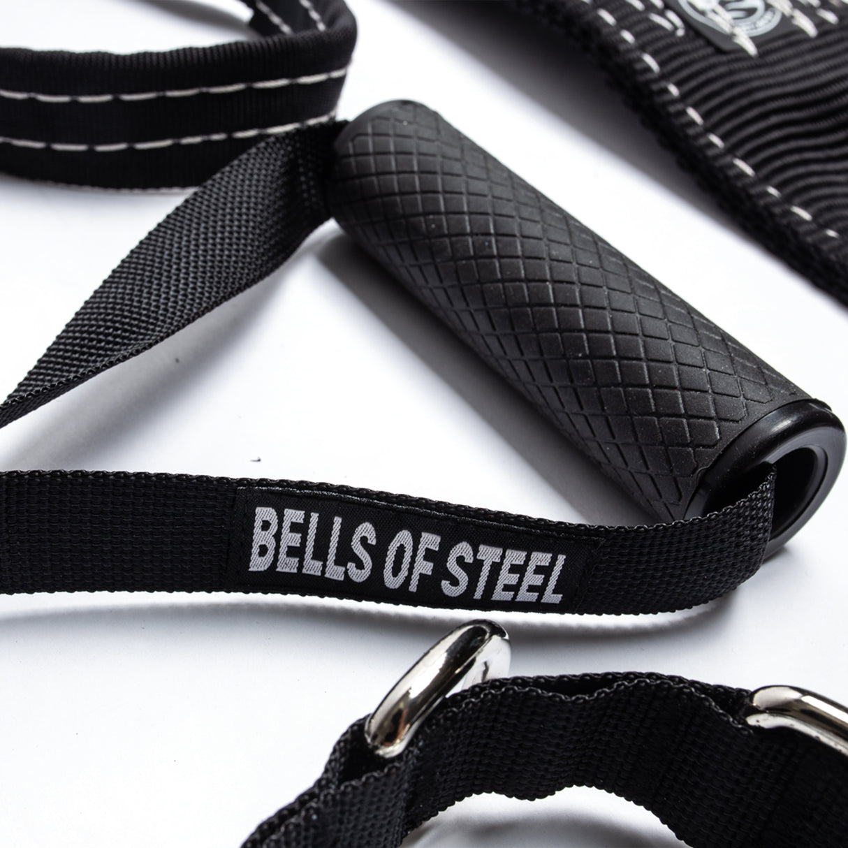 Close-up of a fitness strap with a black, textured handle reading "BELLS OF STEEL" in white, from the Suspension Spotter Straps And Bodyweight System. The handle connects to a black cord with silver clips, all set against a white background.