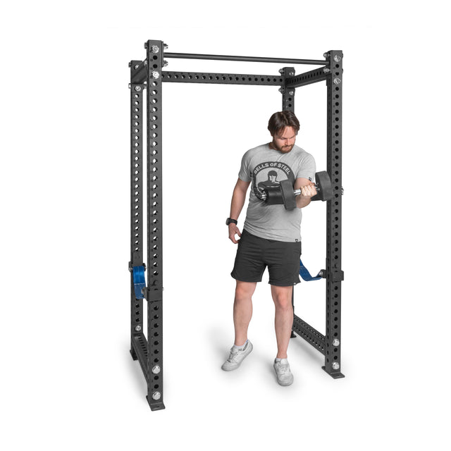 A man in a gray shirt and black shorts performs a bicep curl with a dumbbell inside a Bells of Steel rack featuring the Split Squat Leg Roller Rack Attachment. The black metal rack offers multiple holes for adjustability and includes a pull-up bar at the top.