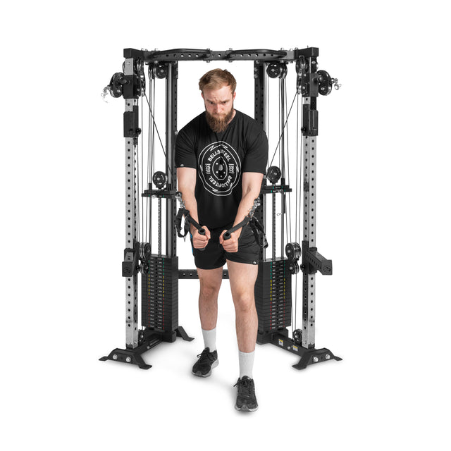 A bearded man in a black T-shirt and shorts exercises on the Bells of Steel Functional Trainer Cable Tower, efficiently pulling down weights with both hands using its adjustable pulleys and weight stacks.