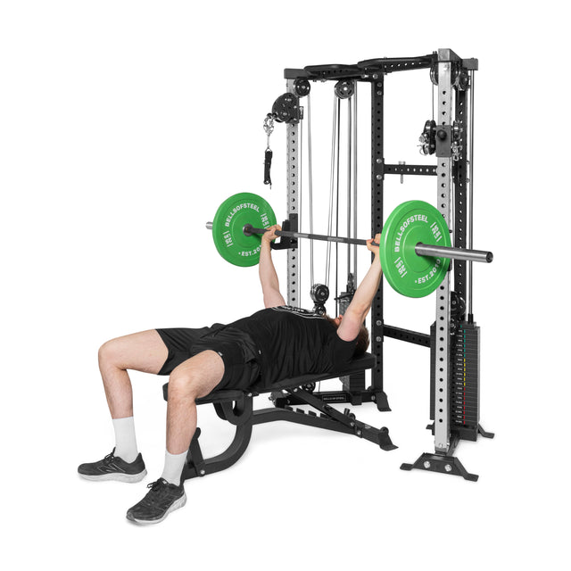 Dressed in athletic wear, a person on a bench uses the Bells of Steel Functional Trainer Cable Tower with green weight plates to optimize their workout with its various pulleys and cables.