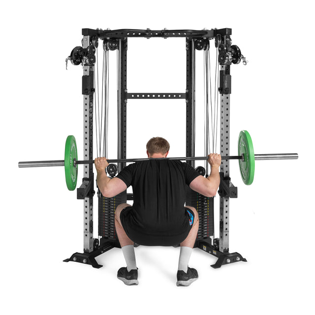 A person with short brown hair is squatting with a barbell on their shoulders inside a power rack, surrounded by Bells of Steel's Functional Trainer Cable Tower. The barbell has green weight plates, complementing their black attire and shoes, enhancing their functional trainer routine.