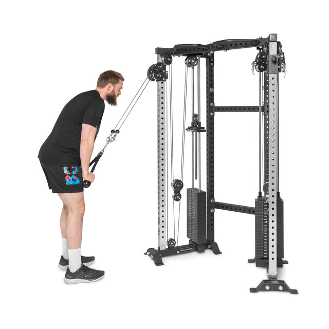 A person in a black shirt and shorts uses the Bells of Steel Functional Trainer Cable Tower for tricep exercises, gripping the handles with weights stacked, set against a plain white background.