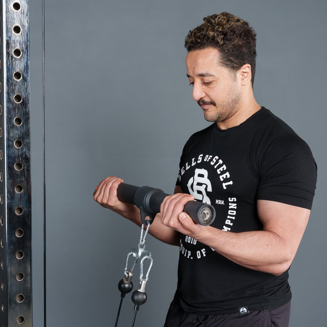 A man in a black "Bells of Steel" shirt performs a cable bicep curl with the Bells of Steel Fat Bar - Straight Handle Bar on a cable machine in a gym.