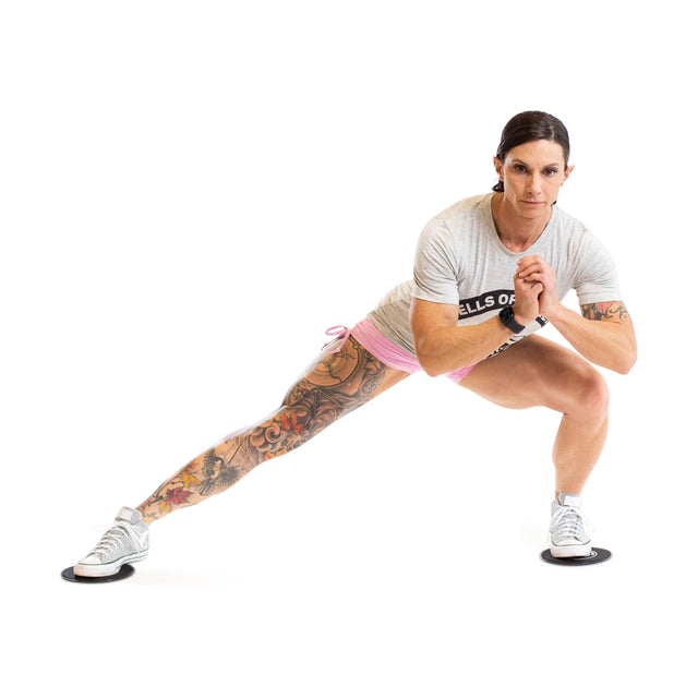A person with tattoos on one leg performs a lateral lunge with Bells of Steel Core Sliders, wearing a gray t-shirt, pink shorts, and sneakers, hands clasped in front of their chest on a white background.