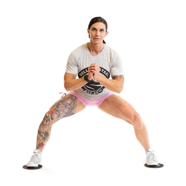 A woman with a large left leg tattoo performs a side lunge using Bells of Steel Core Sliders, hands clasped at her chest, demonstrating their use in a total body workout. She wears a gray t-shirt and pink shorts.