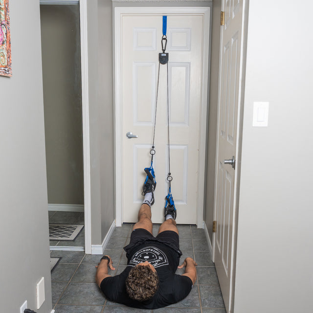 A person lies face up on the floor in a home hallway, performing a bodyweight workout with both feet secured in the Bells of Steel Rotational Pulley system, which is attached to the top of a closed door.
