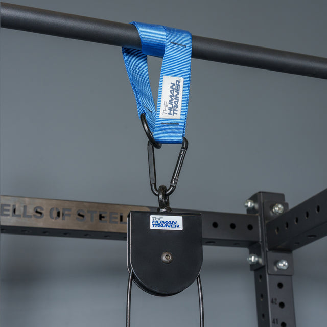 A blue strap labeled “The Human Trainer” hangs from a pull-up bar, attached to the Bells of Steel Rotational Pulley kit. The setup is part of a metal workout rack, designed for effective bodyweight workouts and shown against a gray background.