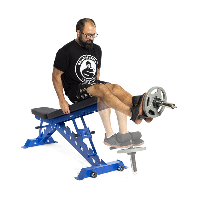 A man is seated on a blue exercise bench, doing leg extensions with knee protection. He grips the sides and utilizes the Bells of Steel Canada Tibialis Trainer Bar, loaded with Olympic weight plates attached to his legs. His attire consists of a black t-shirt and shorts.