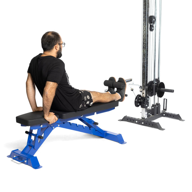 A man in a black shirt and shorts sits on a blue bench, using the Bells of Steel Tibialis Trainer Bar with Olympic plates, his legs extended and feet on the padded attachment to help prevent knee injuries.