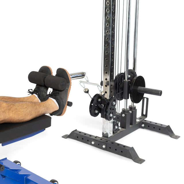 A person wearing black athletic shoes is utilizing a Tibialis Trainer Bar from Bells of Steel Canada at the gym. This equipment includes Olympic weight plates for added resistance and features a pulley system along with padded rollers to protect the knees during exercise.