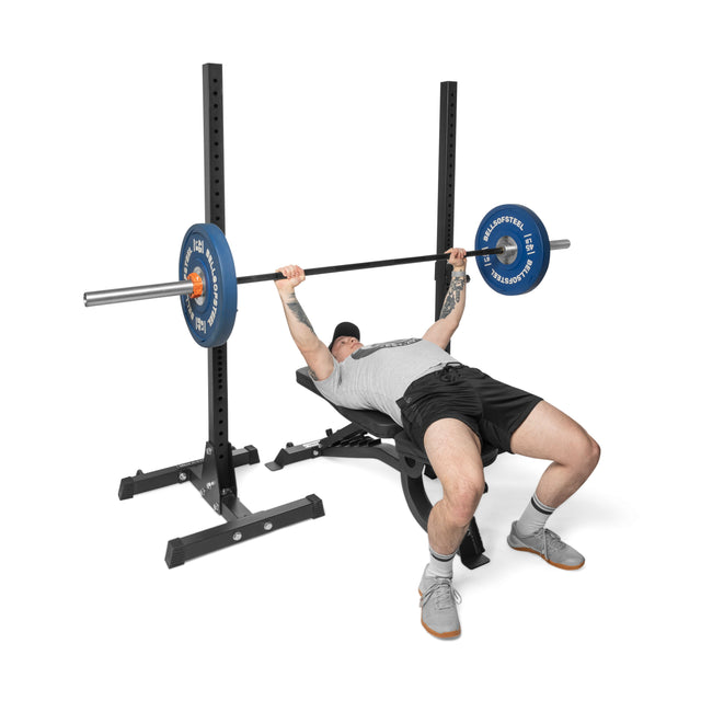 A person in a gray shirt and black shorts performs a bench press with blue weight plates on the Bells of Steel Adjustable Utility Bench, which has a 575lb max capacity.