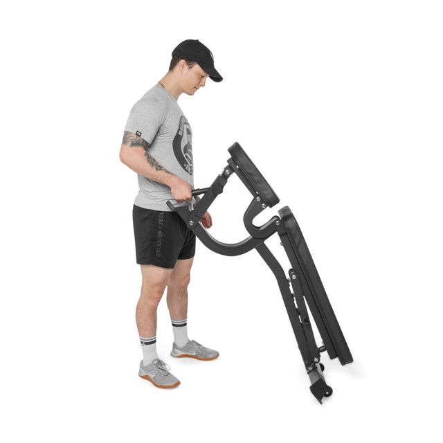 A man in athletic wear and a cap stands on a white background, lifting one end of the Bells of Steel Adjustable Utility Bench to display its versatile incline angles and included bench attachments.