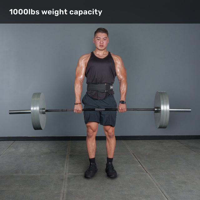 A muscular man in a gym holds the Bells of Steel Multi-Purpose Olympic Barbell – The Utility Bar, wearing a black tank top, shorts, and a weightlifting belt. Text above reads “1000lbs weight capacity.”.