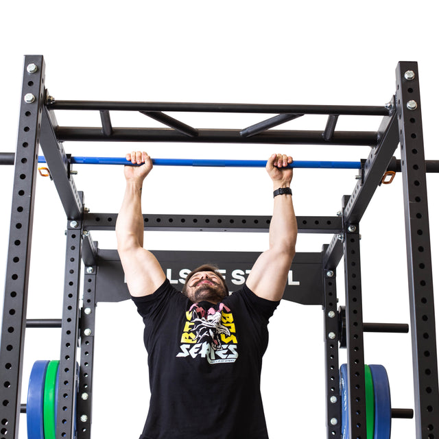 A man in a black graphic t-shirt hangs from the Bells of Steel Above Rack Barbell Holder, attached to a squat rack with weights, preparing for a pull-up against a white background.
