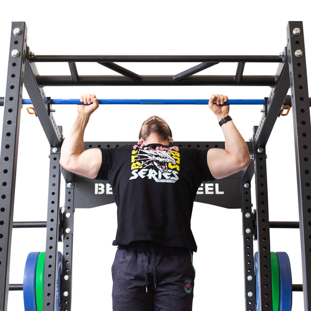 A person uses the Bells of Steel Above Rack Barbell Holder, attached to a black metal rack, performing a pull-up. They wear a black T-shirt with a colorful graphic and black pants. The scene has a plain white background.