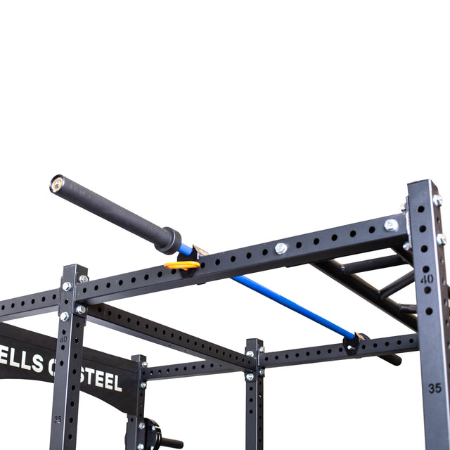 A close-up of the Bells of Steel Above Rack Barbell Holder in black and blue, attached to a metal rack with adjustable holes and ladder-style bars, with part of the "Bells of Steel" logo visible in the background.