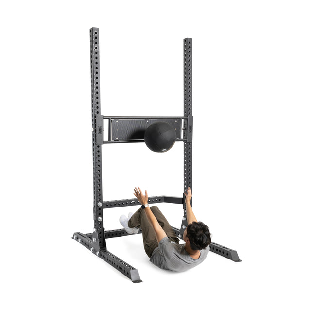 Using the Bells of Steel Utility Seat for Hydra/Manticore, a person performs sit-ups and throws a medicine ball at a metal rebounder rack in a gym space designed for wall ball workouts.
