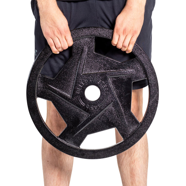 A person grips a Black Mighty Grip Olympic Weight Plate set by Bells of Steel. With both hands on its precision handles, they hold the star-patterned plate in front of their legs. Clad in black shorts, their legs are partially visible beneath.