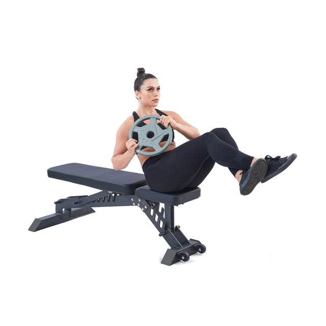In a white setting, a woman in black workout gear performs ab exercises on a bench with her feet off the ground. She twists her upper body while holding a Gray Mighty Grip Olympic Weight Plate by Bells of Steel, keeping her bun neat and tidy.