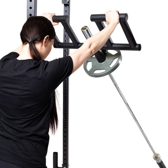 A person with long dark hair in black workout gear grips the handles of a Bells of Steel Viking Press Attachment connected to an Olympic bar loaded with weight plates, preparing for exercise.