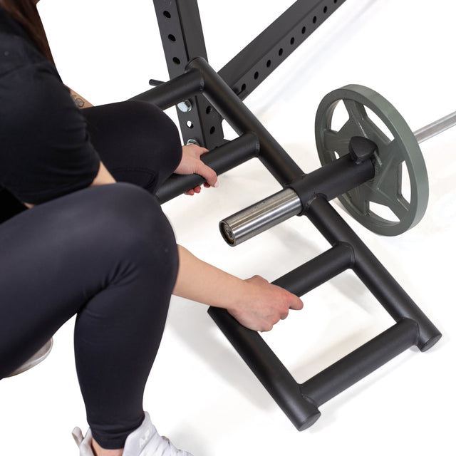 Wearing black leggings and white sneakers, a person grips the Bells of Steel Viking Press Attachment connected to a 2" Olympic bar, getting ready for a landmine exercise in the gym.