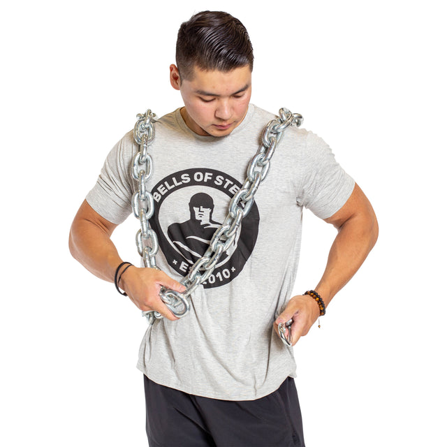 A man in a gray Bells of Steel t-shirt and black shorts stands with the Chain Weight Vest draped over his shoulders, gripping it with both hands and looking down, ready for intense strength training.