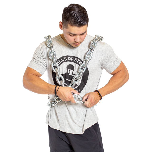 A man wears the Bells of Steel Chain Weight Vest over a light gray t-shirt and black shorts, adjusting the vest as he looks down. The white background emphasizes his strength training focus.