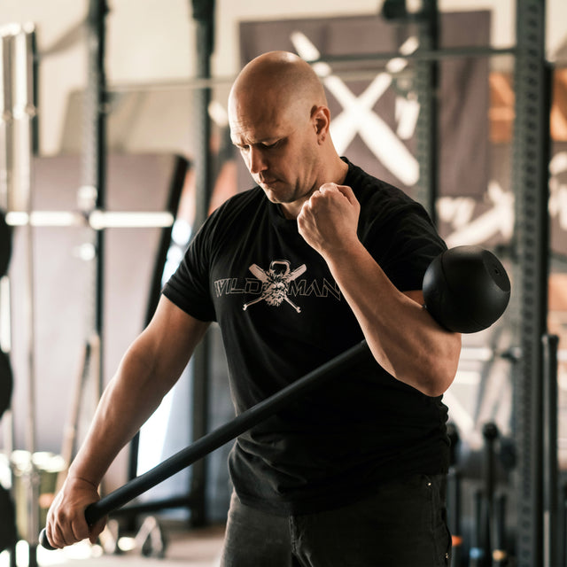 A bald man in the gym swings a Bells of Steel Adjustable Macebell with both hands, boosting core strength. Focused and intense in a black skull T-shirt, weights and racks set the backdrop for his rigorous workout.