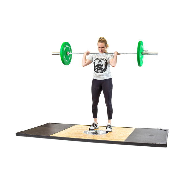 A woman stands on a weightlifting platform, holding the Bells of Steel Oly Lifting Needle Bearing Bar - Women's with green plates (25mm shaft) across her upper back, preparing to lift. She wears a light t-shirt, black leggings, and athletic shoes.