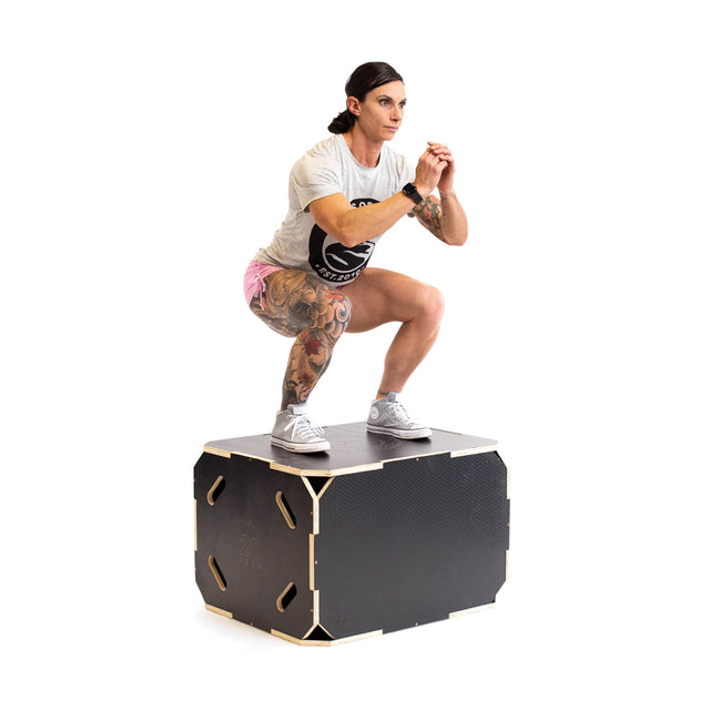 A person with tattoos squats on a Bells of Steel 3 in 1 Anti-Slip Wood Plyo Box, wearing a T-shirt, pink shorts, and sneakers—ideal for any home gym against a white background.