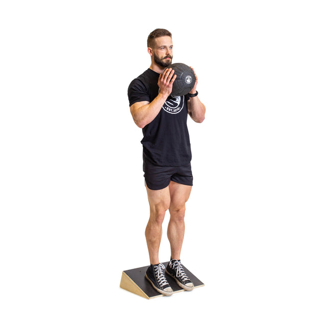 A man in black workout clothes prepares for exercise by holding a weight plate at chest level while standing on a Bells of Steel Squat Wedge Board, with a plain white background highlighting his focus on improving squat depth.