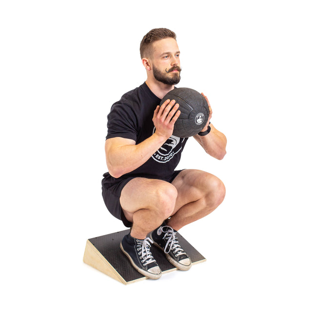 A bearded man wearing a black t-shirt and shorts squats on the Bells of Steel Squat Wedge Board while holding a medicine ball, demonstrating how it enhances ankle mobility and squat depth.
