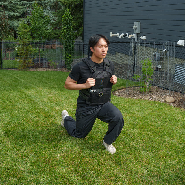 Wearing the Bells of Steel Ultimate Backyard Training Bundle, a person dressed in black workout clothes performs lunges on a grassy lawn in their fenced backyard, maximizing their outdoor workout with this essential fitness equipment.