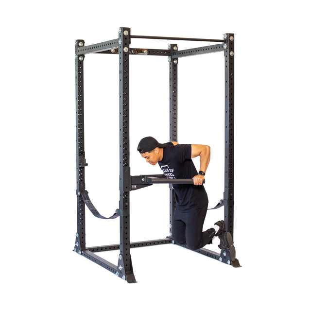 A person in workout attire and a black cap uses resistance bands with the Bells of Steel Y Dip Bar Rack Attachment inside a black power rack. They kneel on the bands, gripping the rack's handles, while the sturdy structure with adjustable heights is protected by UHMW lining.