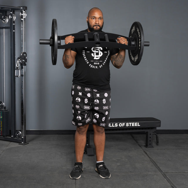 A man in black workout clothes stands in a gym, holding the Arch Nemesis Tricep Bar by Bells of Steel across his chest near a weight bench and strength training equipment, facing forward.