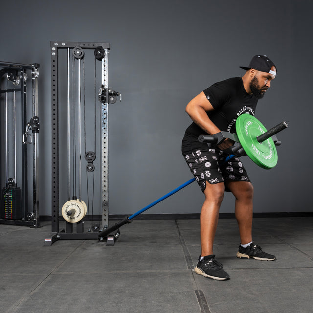 A man in a black T-shirt, shorts, and backward cap performs a landmine row using the Bells of Steel Arch Nemesis Tricep Bar with a barbell and green plate in a gym, with various gym equipment and cables visible in the background.